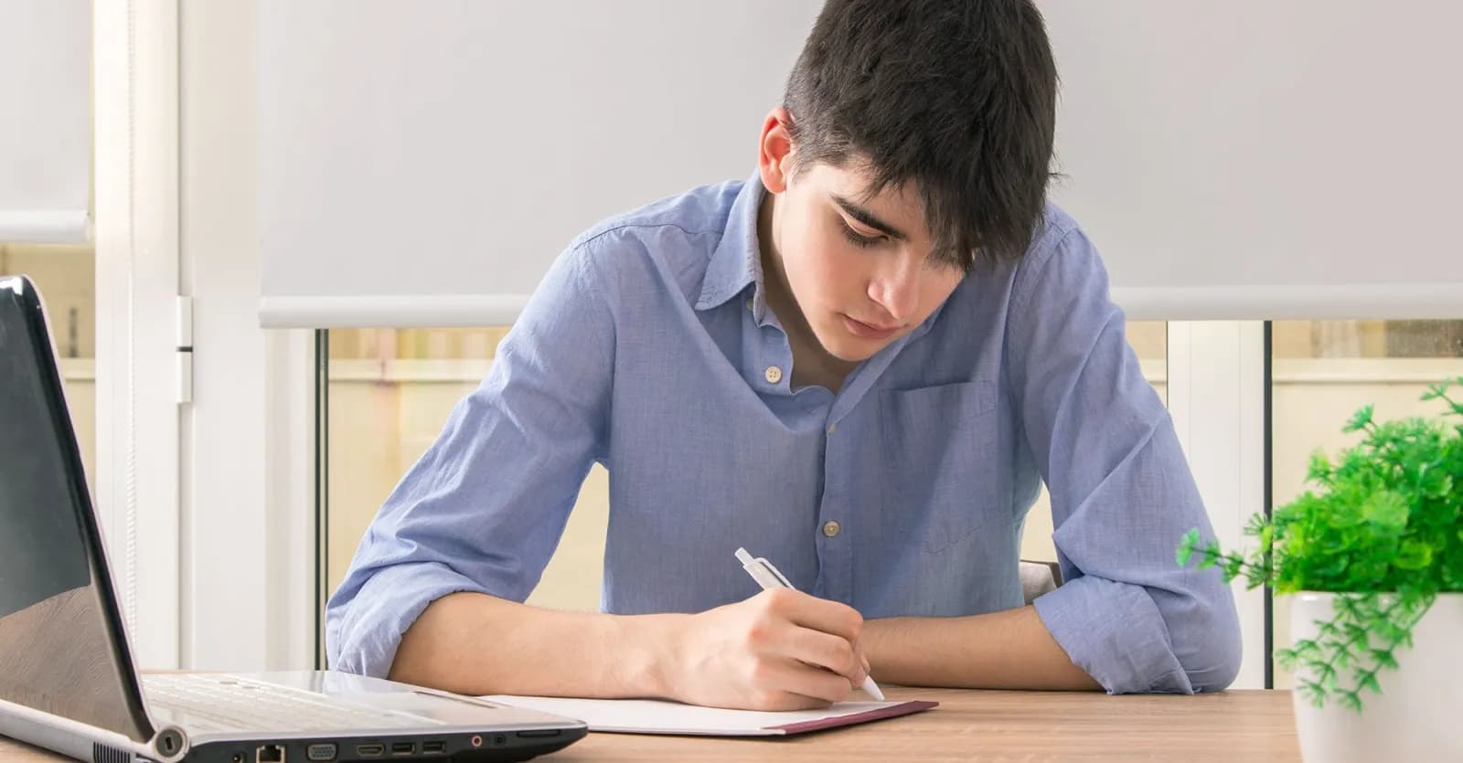 a male Pathfinder special education student sitting at a desk writing in a notebook, a laptop is next to him.